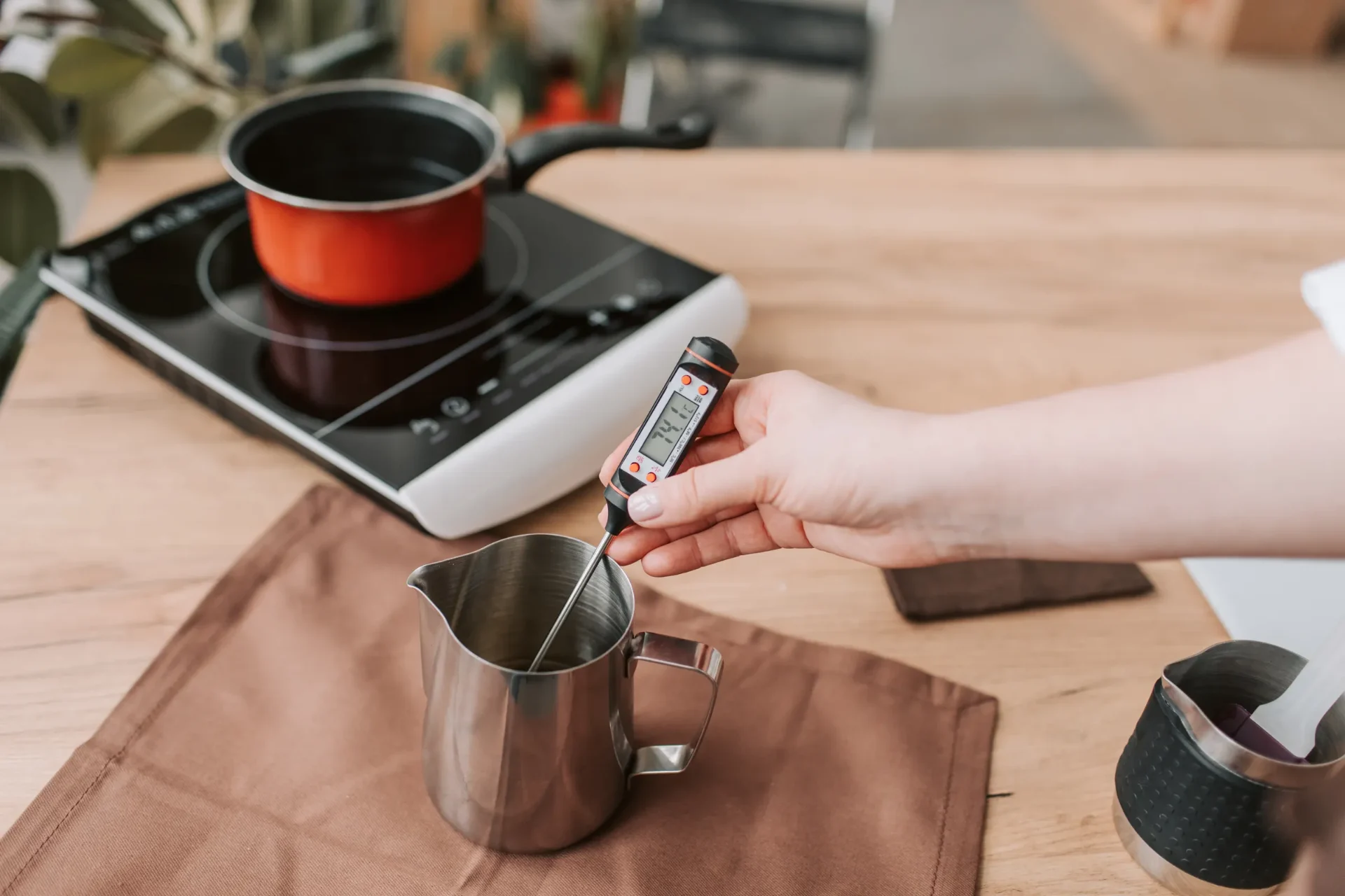 A hand pours syrup into a metal pitcher on a table next to a stovetop and cup.