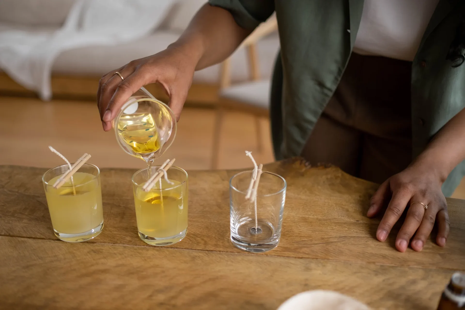 A person is pouring wax into small glass candle moulds with wicks.
