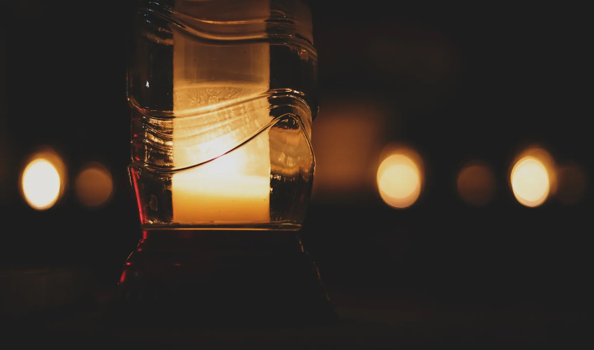 A glass lantern with a lit candle inside sits in front of blurred candlelight.